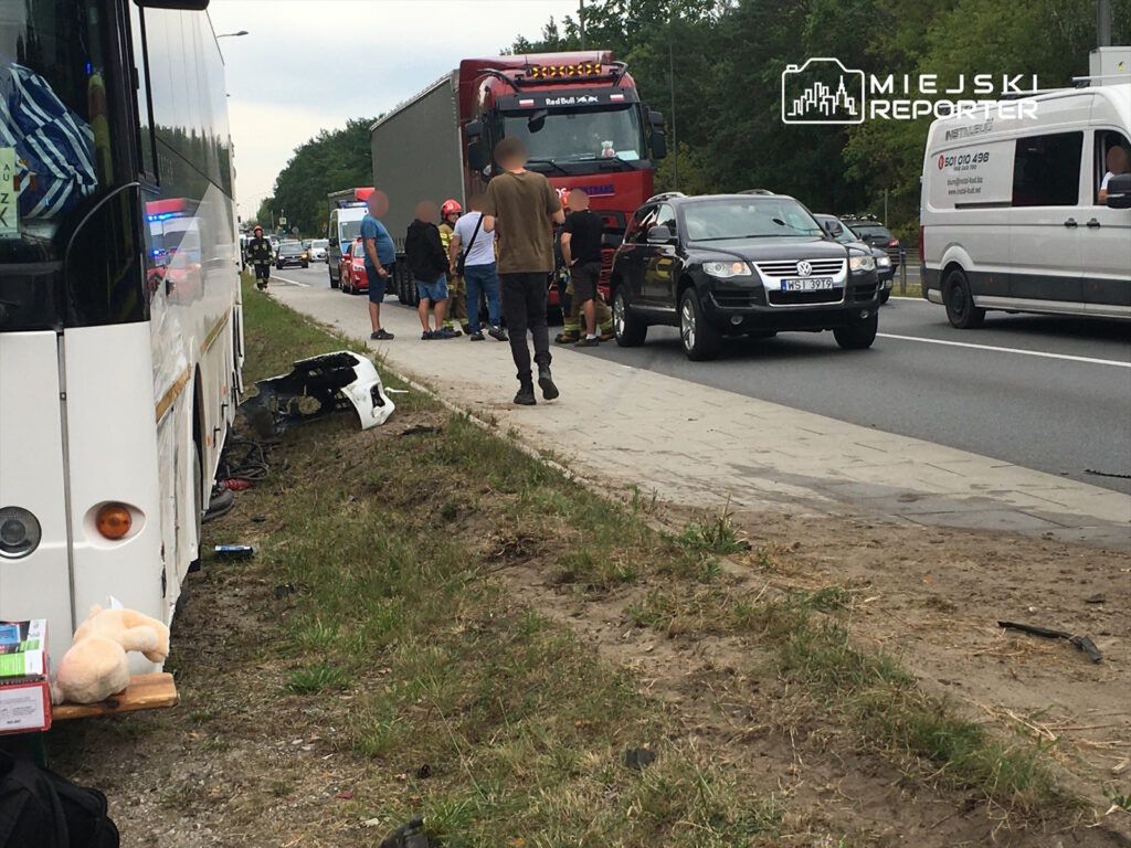 Grupa osób wzywa pomoc przy uszkodzonym pojeździe na poboczu drogi, obok stojących samochodów.