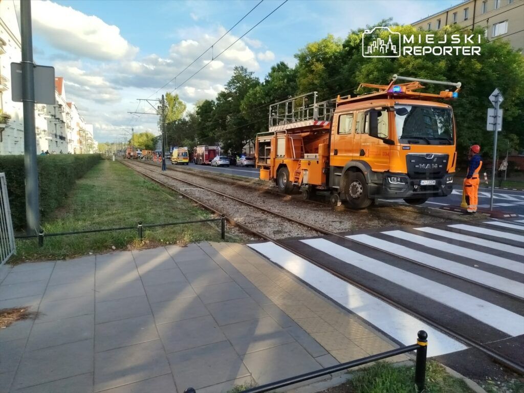 Pomarańczowy pojazd roboczy z podnośnikiem stoi na torach tramwajowych, obok pracowników w odzieży ochronnej.