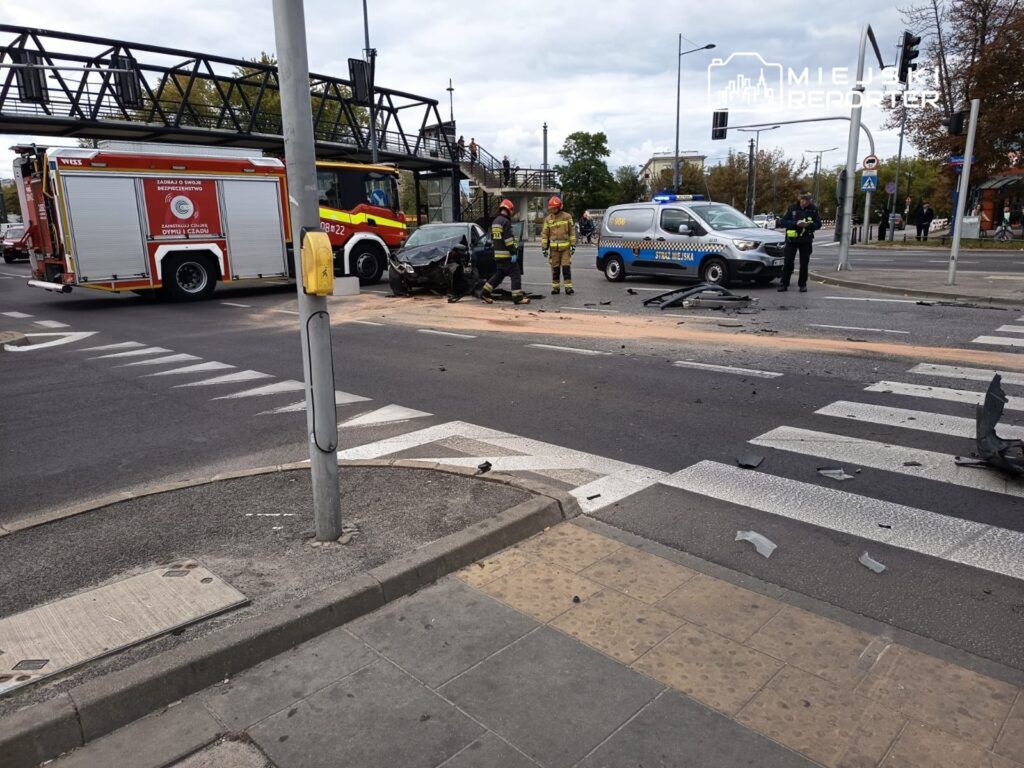 Strażacy oraz policjanci pracują przy miejscu wypadku drogowym, gdzie uszkodzony samochód leży na jezdni.