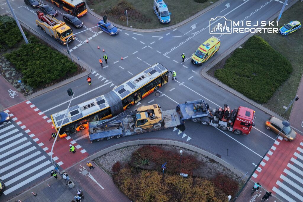 Zablokowany ruch na skrzyżowaniu, gdzie autobus i ciężarówka z maszyną tworzą przeszkodę, a pracownicy drogowi kierują ruchem.