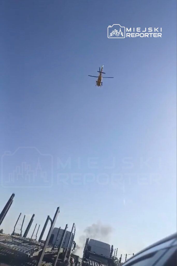 Śmigłowiec przelatujący nad parkingiem z ciężarówkami, w tle widoczny dym.