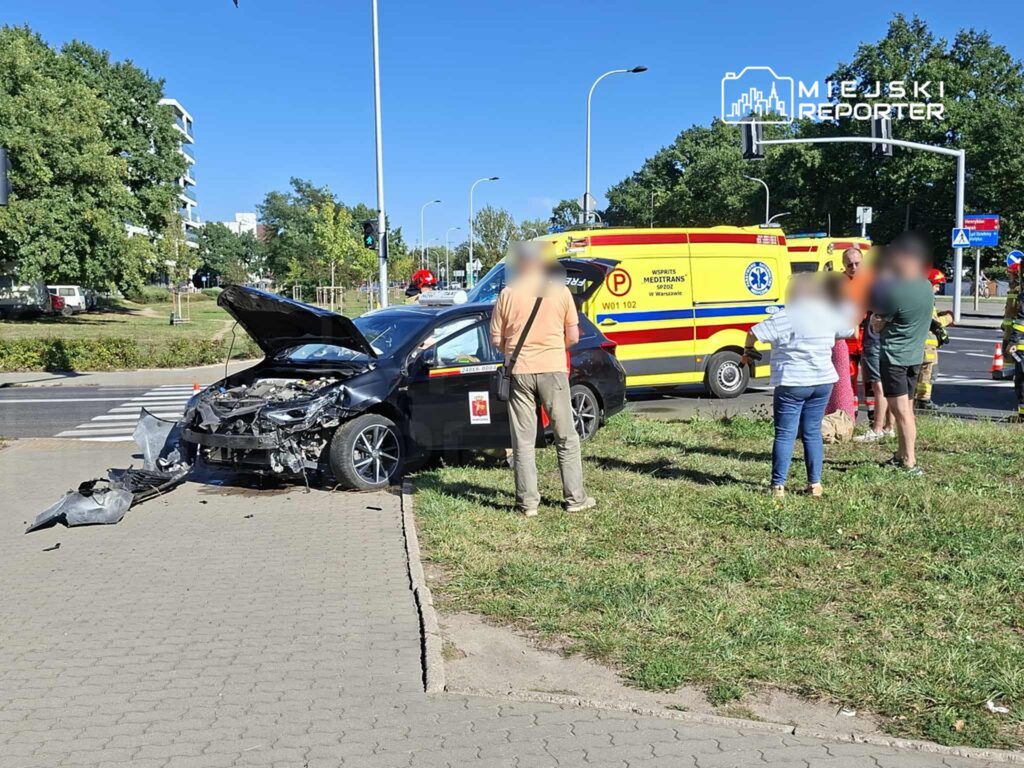 Czarny samochód osobowy z uszkodzoną przednią częścią stoi na chodniku, obok zebra gromadzi się grupa ludzi, w tle widać ambulans.