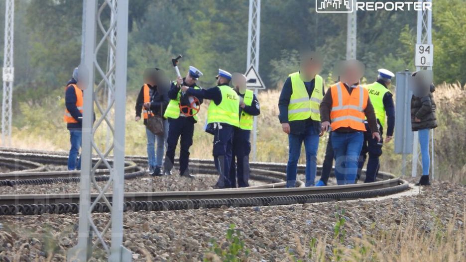 Grupa policjantów i pracowników w odblaskowych kamizelkach prowadzi działania na torach kolejowych w terenie.