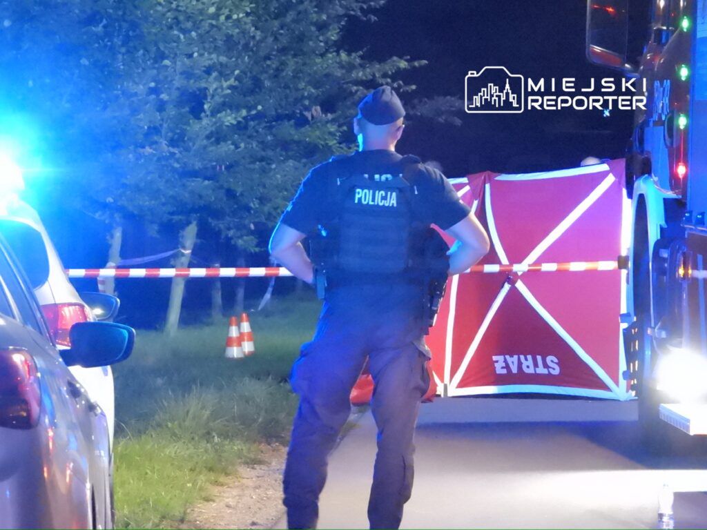 Policjant stoi przy miejscu zdarzenia, zabezpieczonym taśmą i namiotem. W tle widać radiowóz oraz pojazd straży pożarnej. Na miejscu obecne są również światła alarmowe. Sytuacja ma miejsce w nocy, co podkreśla intensywne oświetlenie. Informacje o zdarzeniu są w trakcie zbierania.