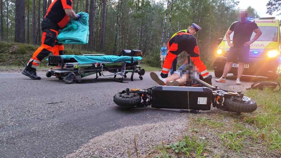 Ratownicy medyczni udzielają pomocy poszkodowanemu na drodze w lesie, obok przewróconego skutera.