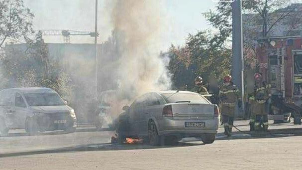 Samochód osobowy wydobywa dym i płomienie, podczas gdy strażacy gaszą pożar na ulicy w mieście.