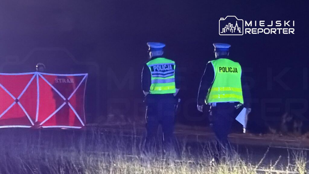 Dwaj policjanci w kamizelkach odblaskowych obserwują miejsce zdarzenia w ciemnej okolicy, obok czerwonej osłony.