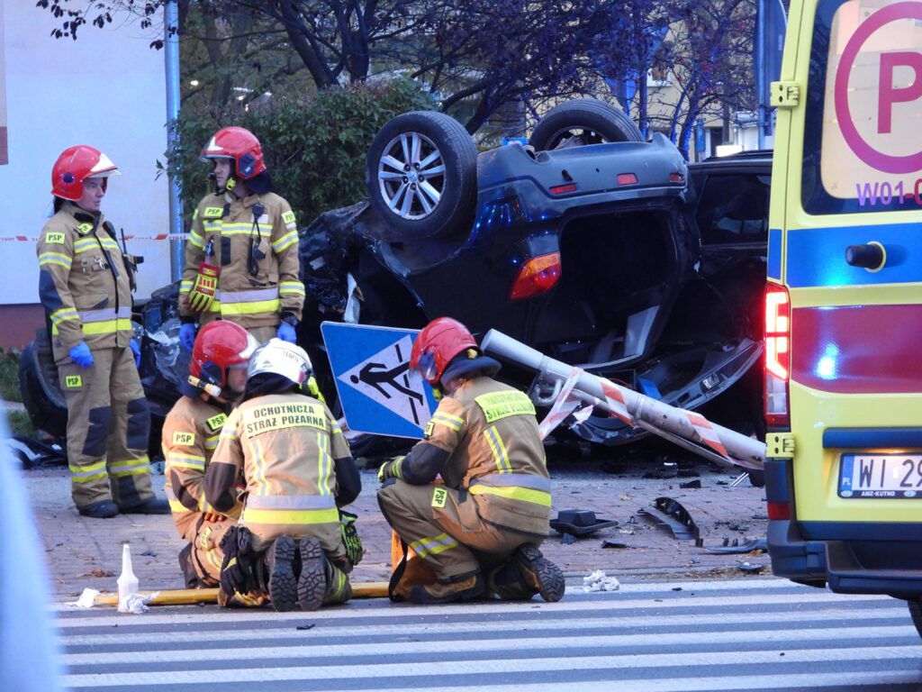 Grupa strażaków w kaskach omawia sytuację obok przewróconego samochodu, który uderzył w znak drogowy.