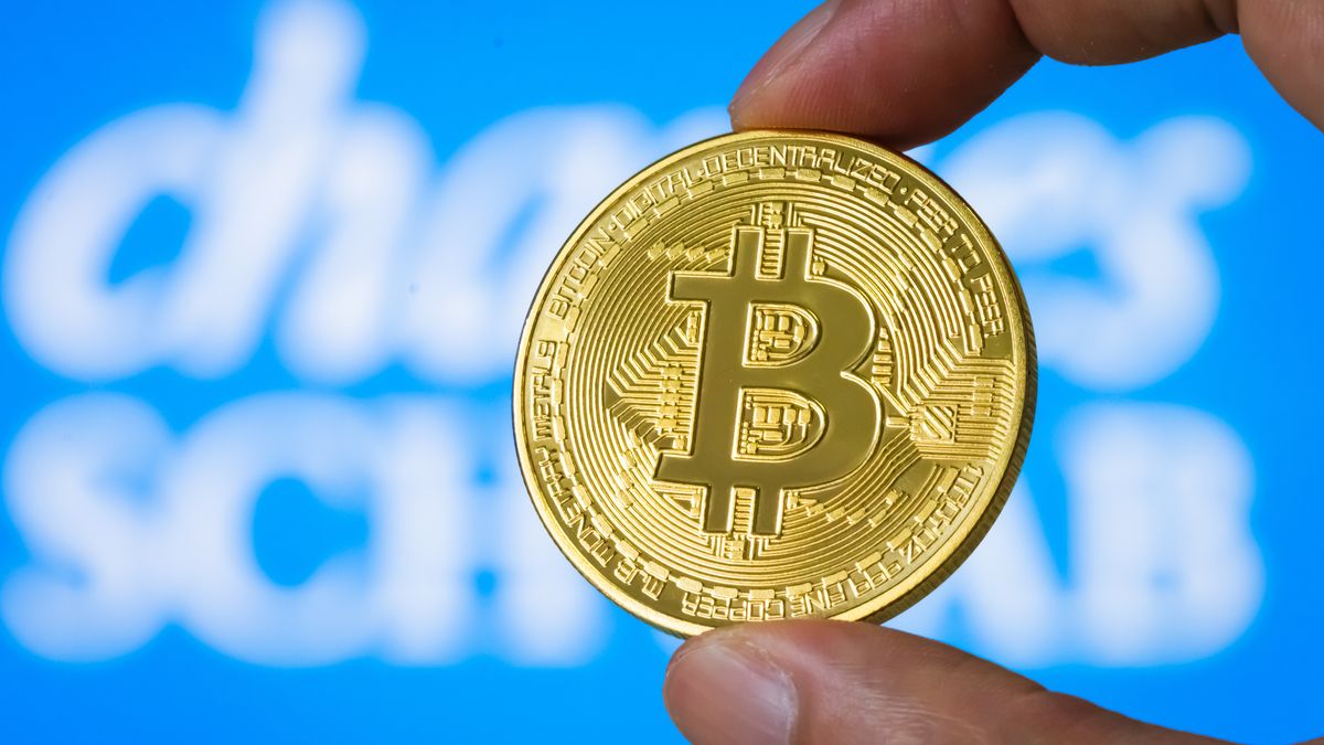 CHONGQING, CHINA - JULY 21: In this photo illustration, a person holds a physical representation of Bitcoin (BTC) in front of a screen displaying the logo of Charles Schwab, symbolizing the growing convergence between digital assets and traditional financial institutions on July 21, 2025 in Chongqing, China. (Photo illustration by Cheng Xin/Getty Images)