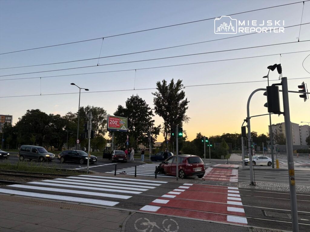 Ruch uliczny na skrzyżowaniu z nowym przejściem dla pieszych, w tle zieleń i billboard reklamowy.