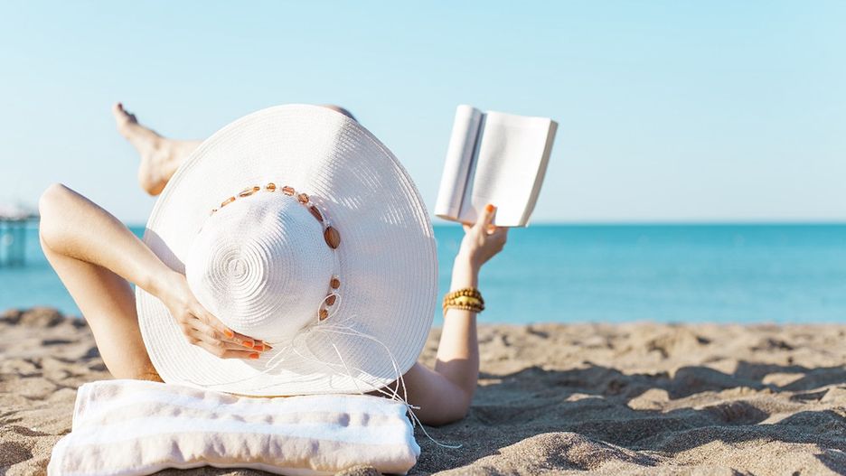 kobieta opala się na plaży w kapeluszu i czyta książkę