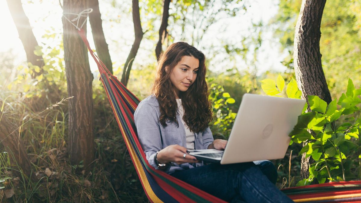 Młoda kobieta pracuje zdalnie, siedząc w hamaku z laptopem.