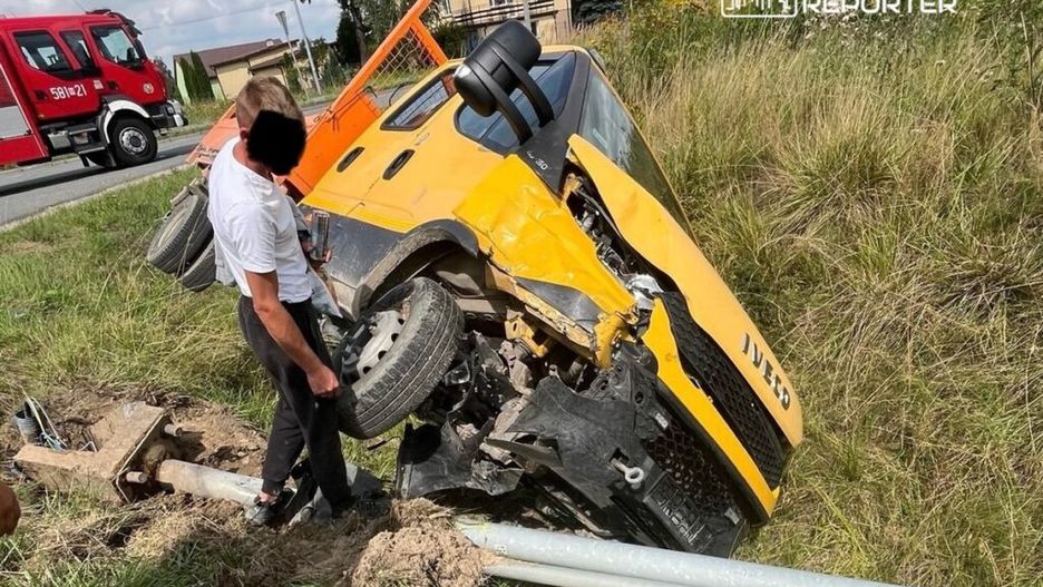 Mężczyzna w białej koszuli sprawdza przewrócony żółty pojazd na poboczu drogi, obok widoczny wóz strażacki.