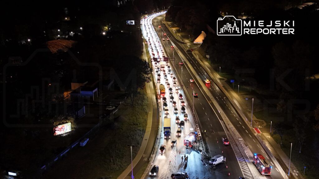 Korki samochodowe na drodze, z pojazdami stojącymi w nocy, w tle widać radiowóz i straż pożarną.