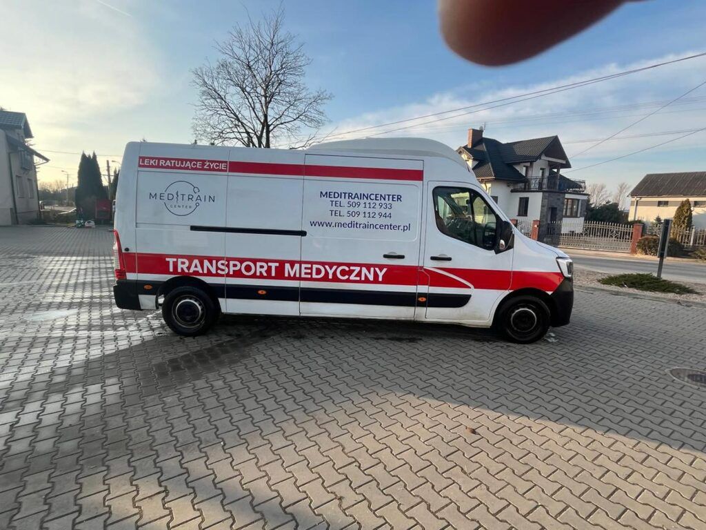 Furgonetka z napisem "TRANSPORT MEDYCZNY" zaparkowana na kostce brukowej w otoczeniu domów.