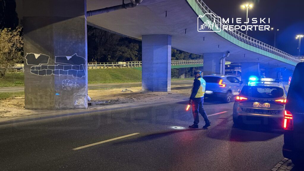 Policjant z latarką kieruje ruchem pod mostem, obok oznakowanego radiowozu policyjnego.