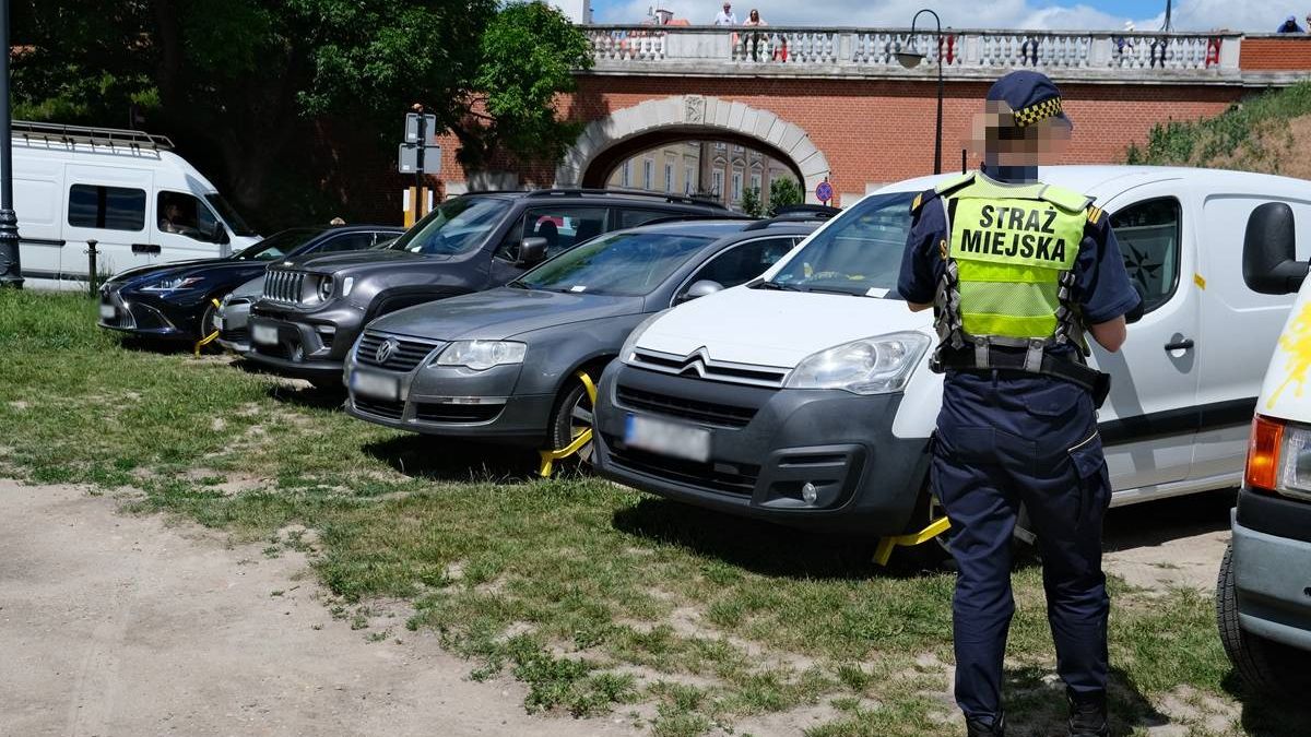 Strażnicy miejscy zakładający blokady na zaparkowane auta na trawniku przy ul. Podwale