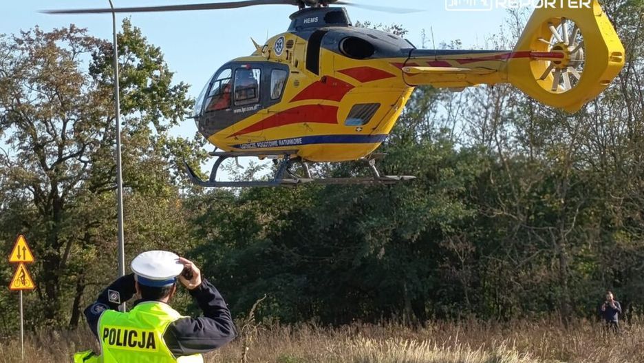 Policjant w żółtej kamizelce fotografuje przelatujący śmigłowiec ratunkowy nad drogą w otoczeniu drzew.
