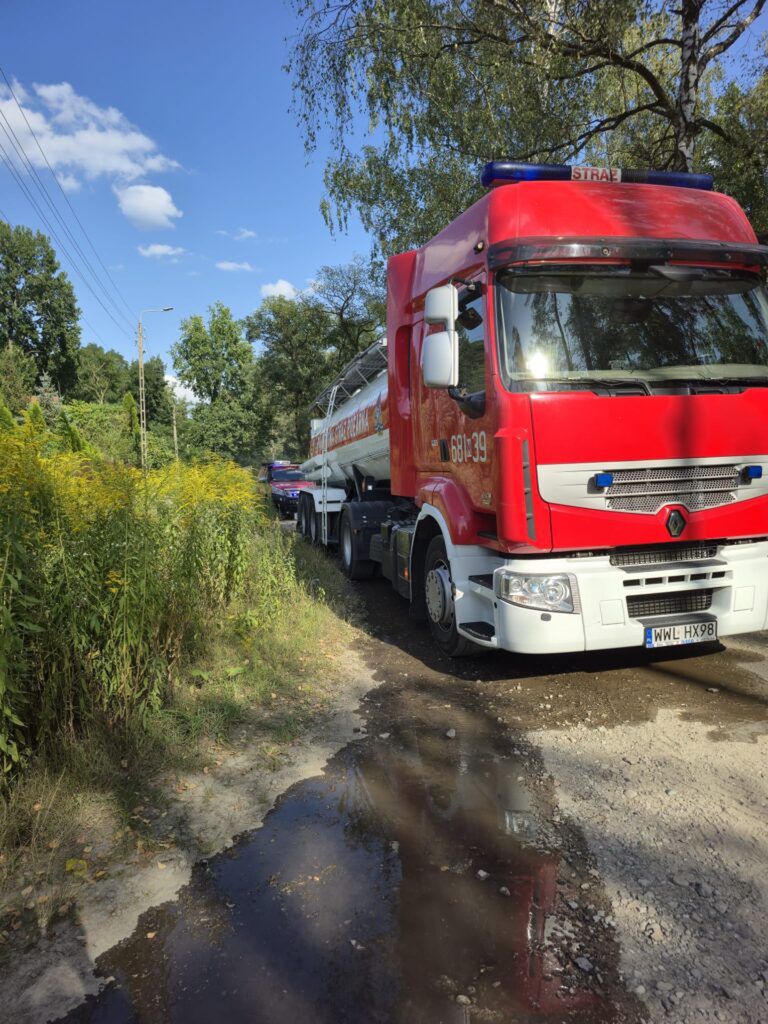 Czerwony ciężarowy samochód strażacki zaparkowany na wąskiej drodze otoczonej zielenią.