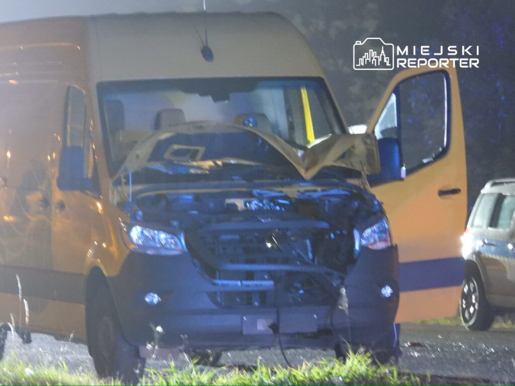 Uszkodzony żółty furgon z otwartą maską stoi na drodze, oświetlony przez niebieskie światła.