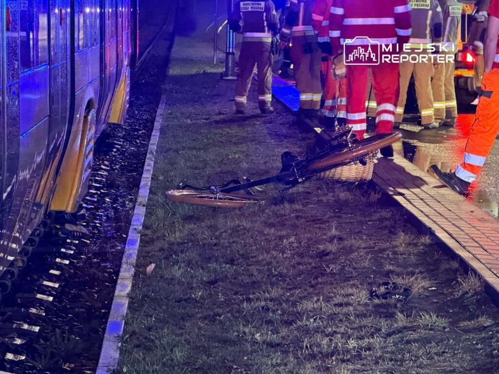 Rower leży na trawie obok torów tramwajowych, w tle znajdują się strażacy w mundurach i pojazdy ratunkowe.