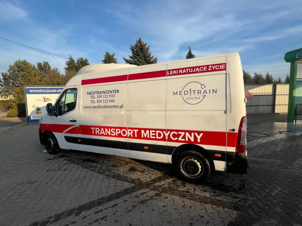 Samochód dostawczy z napisem "TRANSPORT MEDYCZNY" zaparkowany na placu, w tle widoczne drzewa i budynki.