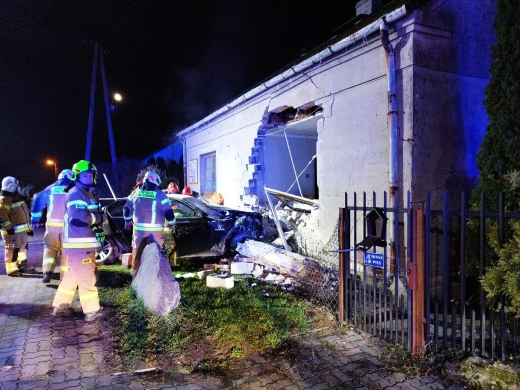 Grupa strażaków w specjalistycznym sprzęcie pracuje przy uszkodzonym budynku po wypadku samochodowym.