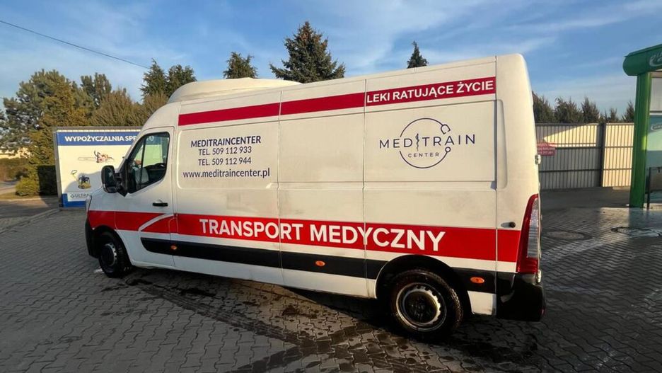 Samochód dostawczy z napisem "TRANSPORT MEDYCZNY" zaparkowany na placu, w tle widoczne drzewa i budynki.