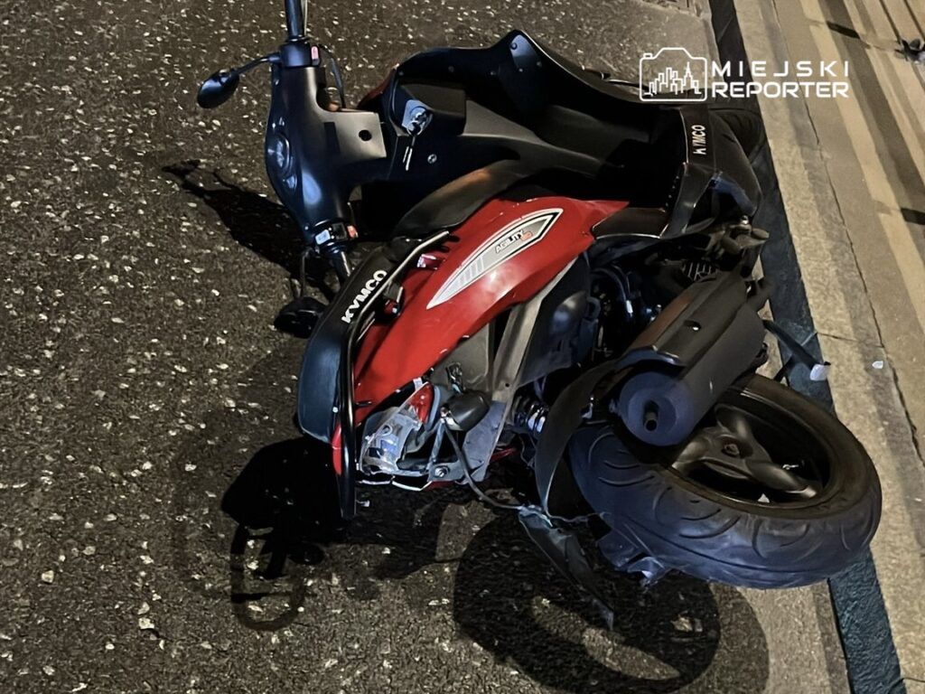 Czerwony skuter leży na boku na asfaltowej drodze w nocy, z widocznymi uszkodzeniami.