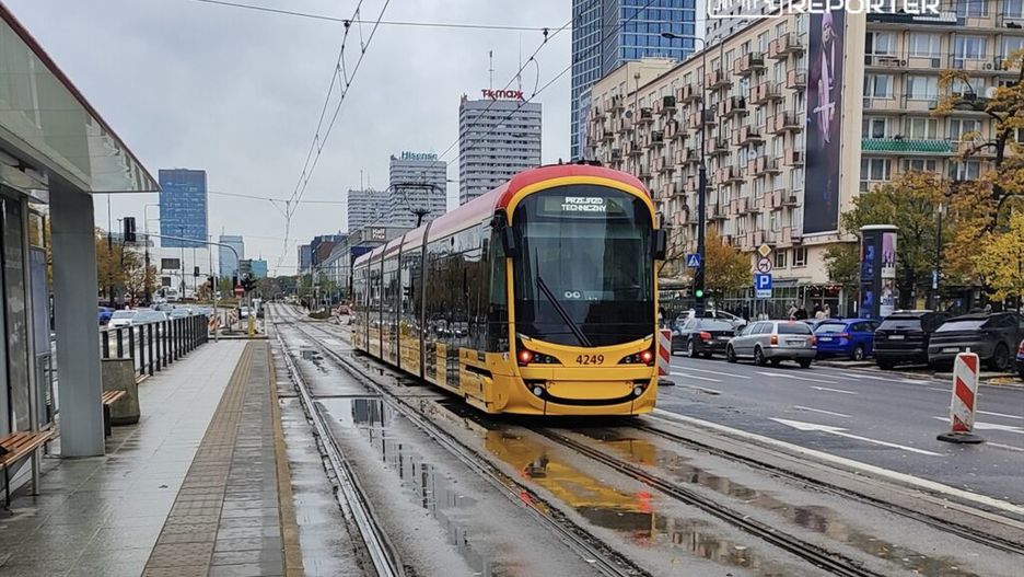 Tramwaj w kolorze żółtym przemieszcza się po mokrej jezdni w miejskim otoczeniu, otoczony nowoczesnymi budynkami.