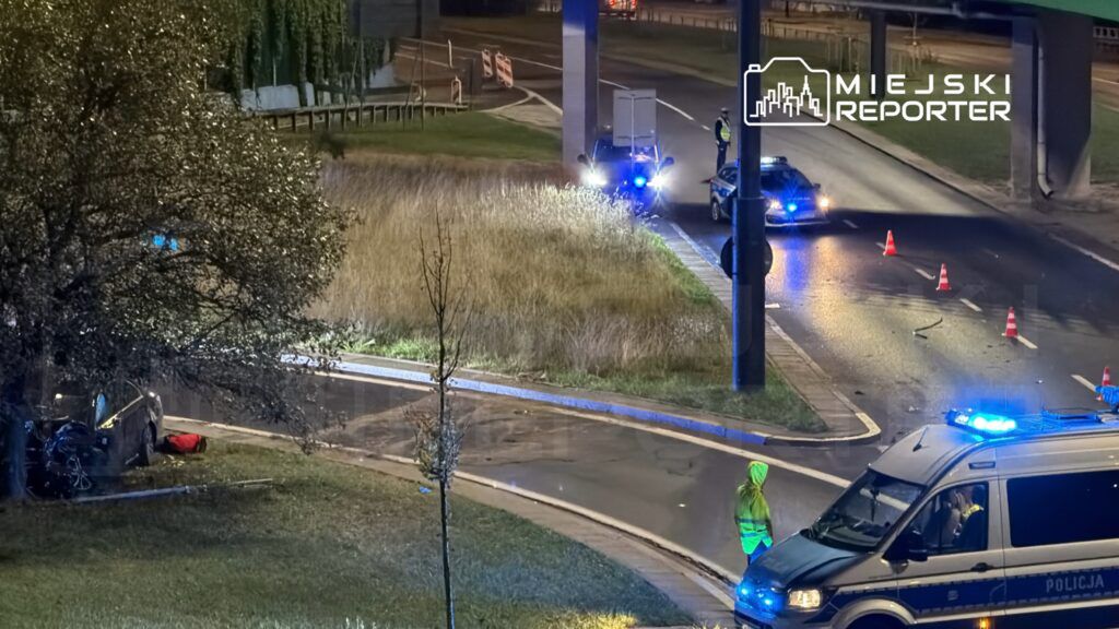 Policyjny radiowóz z włączonymi sygnałami świetlnymi stoi obok miejsca wypadku drogowego, gdzie leży uszkodzony pojazd.