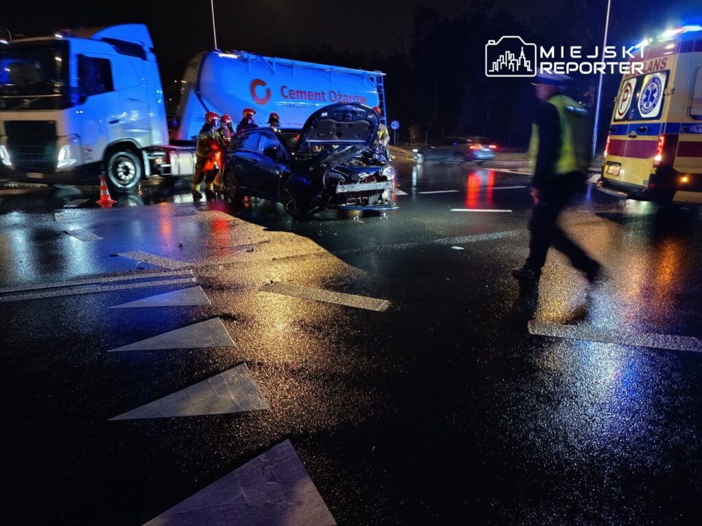 Na mokrej jezdni w nocy strażacy udzielają pomocy poszkodowanemu w wypadku samochodowym, obok stoi ciężarówka.