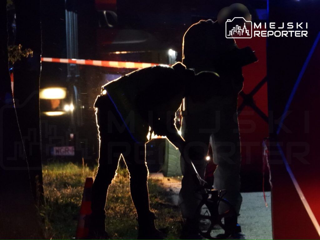 Osoby w mundurach przeprowadzają działania na miejscu zdarzenia w nocy. W tle widoczne są pojazdy służb ratunkowych oraz barierki zabezpieczające teren. Scena oświetlona jest przez reflektory, co podkreśla intensywność sytuacji. Widać także narzędzia używane do analizy miejsca zdarzenia.