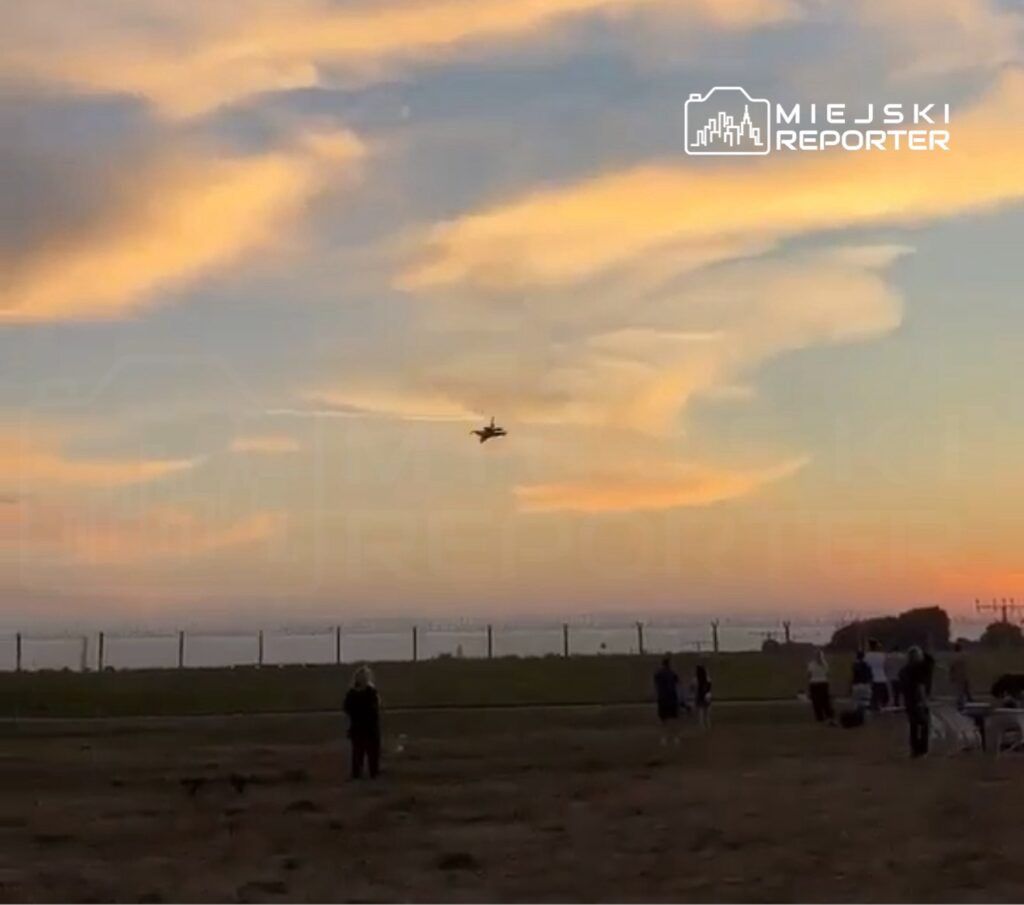 Samolot w locie nad grupą ludzi stojących na otwartym terenie podczas zachodu słońca.