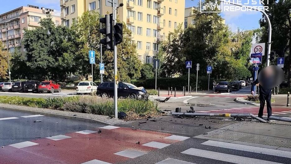 Na skrzyżowaniu zniszczony słup sygnalizacji świetlnej, a obok stoi mężczyzna w czarnym ubraniu, oceniając sytuację.