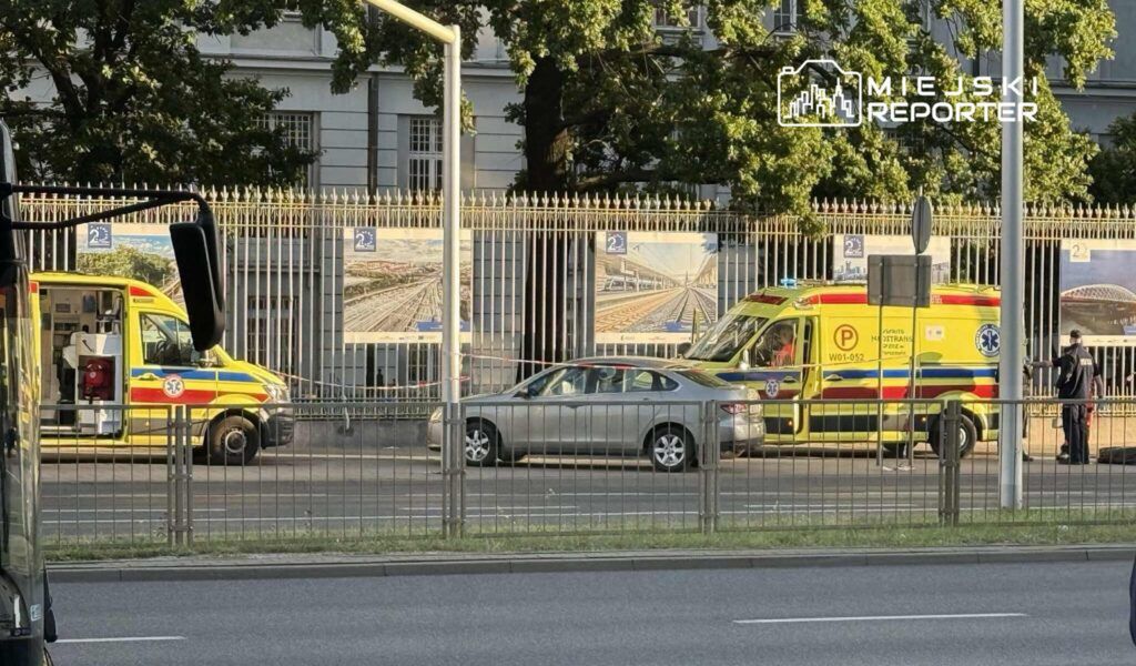 Karetka pogotowia oraz radiowóz stoją na ulicy obok szarego samochodu, a w tle widoczna jest ogrodzona przestrzeń z plakatami.