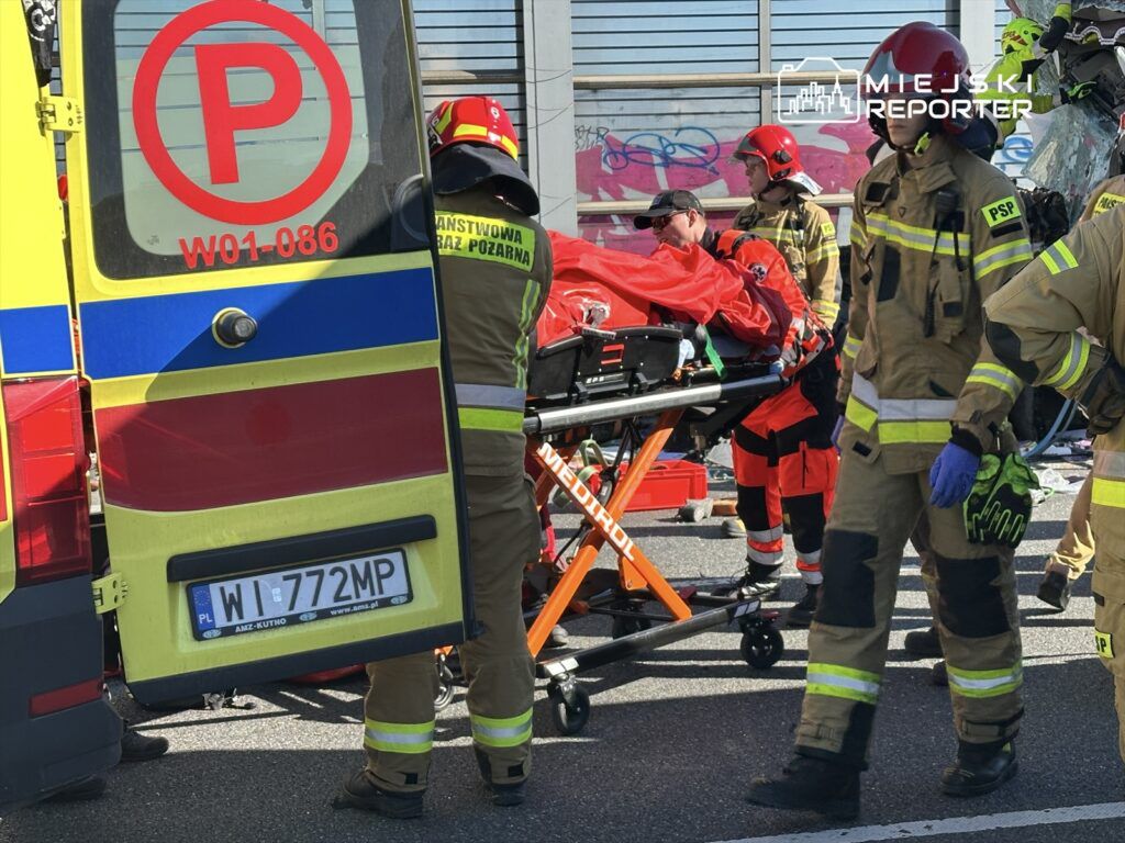 Ratownicy medyczni i strażacy udzielają pomocy poszkodowanemu na noszach w pobliżu karetki.