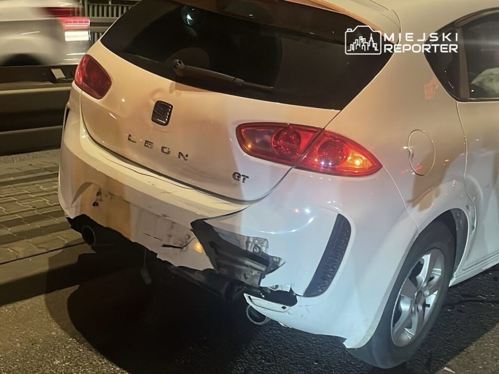 Uszkodzony biały samochód marki Seat Leon z wgniecionym tyłem stoi na drodze w nocy.