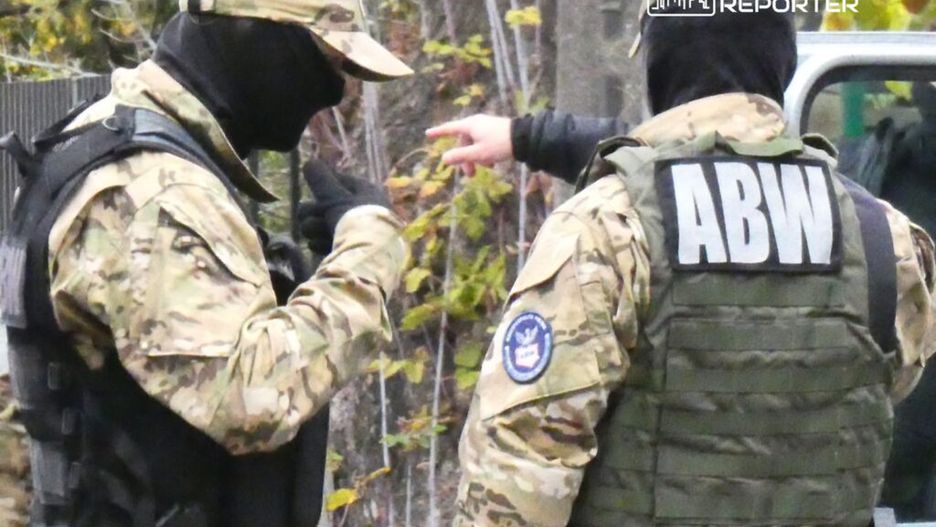 Dwaj funkcjonariusze służb mundurowych w maskach rozmawiają w lesie, jeden wskazuje ręką w kierunku pojazdu.