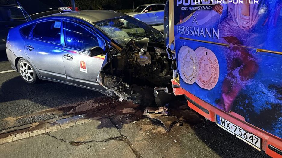 Taksi osobowe z uszkodzoną przednią częścią zderza się z reklamowym autobusem na ulicy, w nocy.