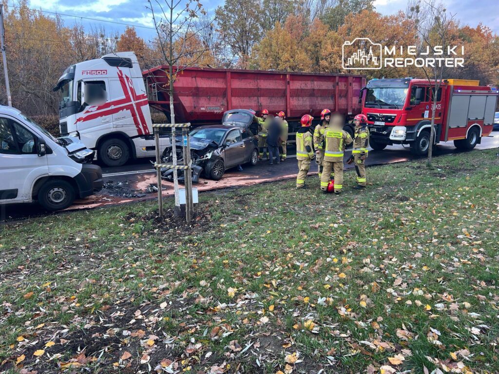Wielu strażaków zabezpiecza miejsce wypadku drogowego z udziałem ciężarówki i osobowego samochodu na drodze otoczonej drzewami.