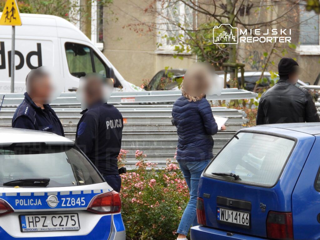 Grupa policjantów rozmawia z osobą w niebieskiej kurtce, stojąc obok metalowych barierek w otoczeniu kwiatów.
