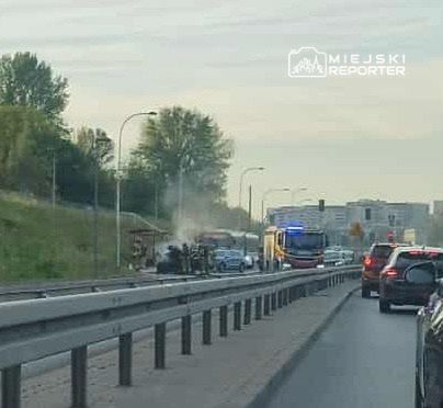 Na drodze widać samochód objęty dymem, obok zatrzymane pojazdy oraz straż pożarną z włączonymi sygnałami świetlnymi.