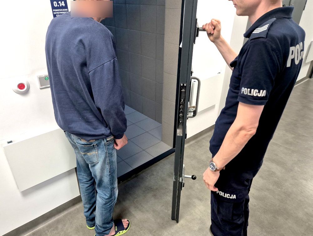 Mężczyzna w dresie stoi w pomieszczeniu, a policjant otwiera drzwi prowadzące do celi. W tle widać szare płytki.