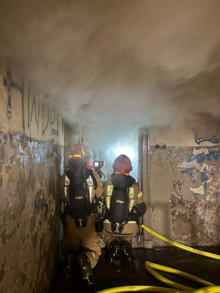 Dwaj strażacy w hełmach i sprzęcie ochronnym, klęczący w zadymionym korytarzu, monitorują sytuację z kamerą.