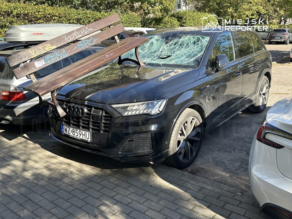 Czarny SUV z uszkodzoną szybą i drewnianą ławką unieruchomioną na dachu, zaparkowany między innymi pojazdami.