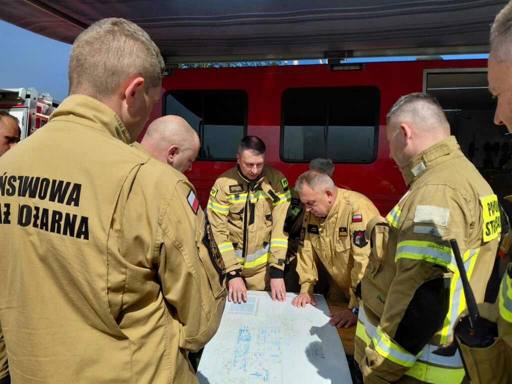 Grupa strażaków w mundurach omawia plan działania przy stole, w tle widoczny wóz strażacki.