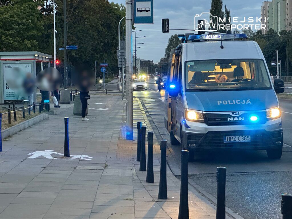 Policyjny radiowóz z włączonymi sygnałami świetlnymi zatrzymany na ulicy, obok przystanku autobusowego.