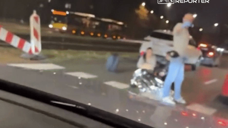 Mężczyzna w niebieskiej koszulce udziela pomocy osobie siedzącej na jezdni, owiniętej w koc termiczny, w nocy.