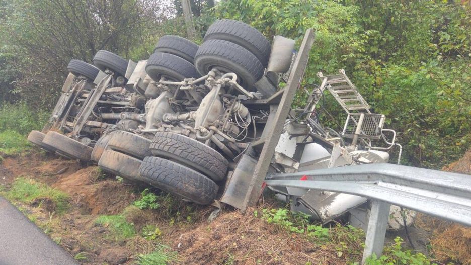 Przewoźnik z przewróconą ciężarówką leżącą na boku w zaroślach przy drodze.